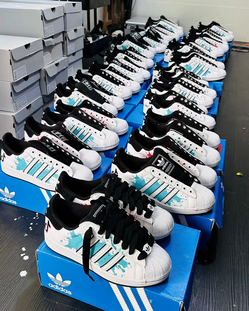 Fileiras de Adidas Superstar pintados à mão nas cores do TikTok, sobre as caixas do pedido