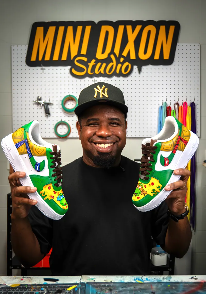 Thiago Peçanha, artista do Mini Dixon Studio, segurando o par de tênis customizado das Tartarugas Ninja diante da placa do estúdio